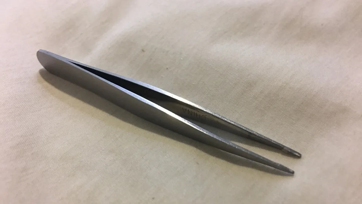 A pair of slanted-tip metal tweezers lying open on a light cloth, the tool most often used to pluck eyebrow hairs