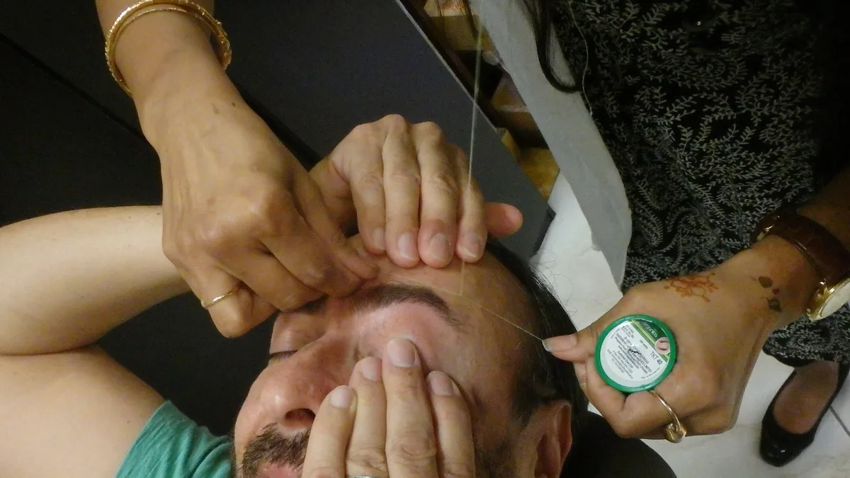 A man lying back with his hands over his eyes while a practitioner threads his eyebrow with cotton thread