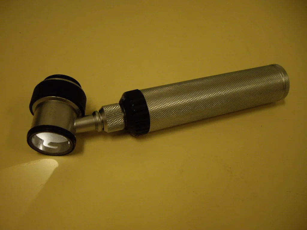 Handheld dermatoscope with a round lens head and a knurled metal handle lying on a table, its built-in light switched on
