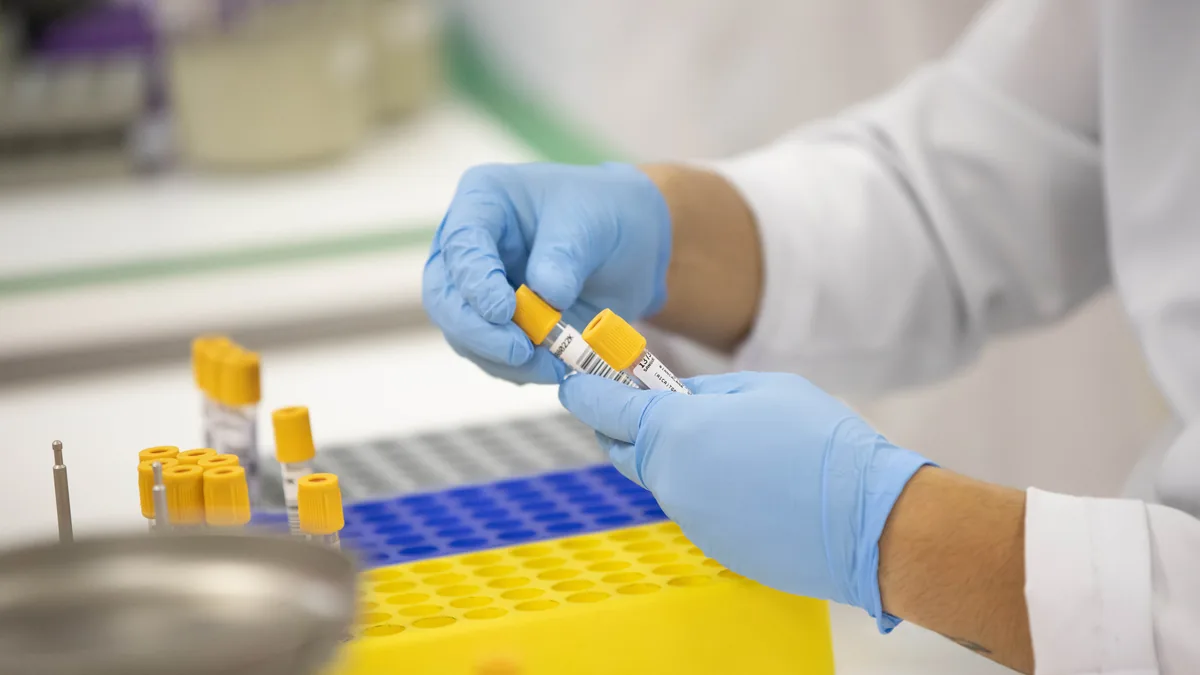 Gloved hands holding two blood sample tubes with yellow caps above a yellow and blue laboratory rack of more tubes