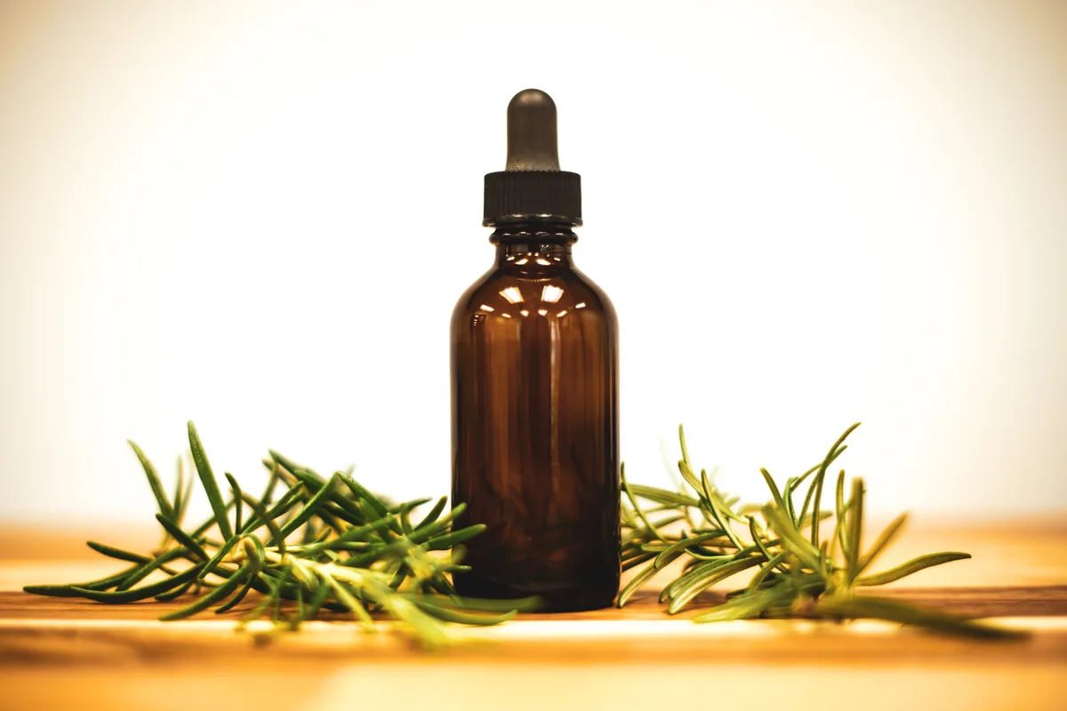 A small glass dropper bottle of rosemary oil lying beside fresh sprigs of rosemary