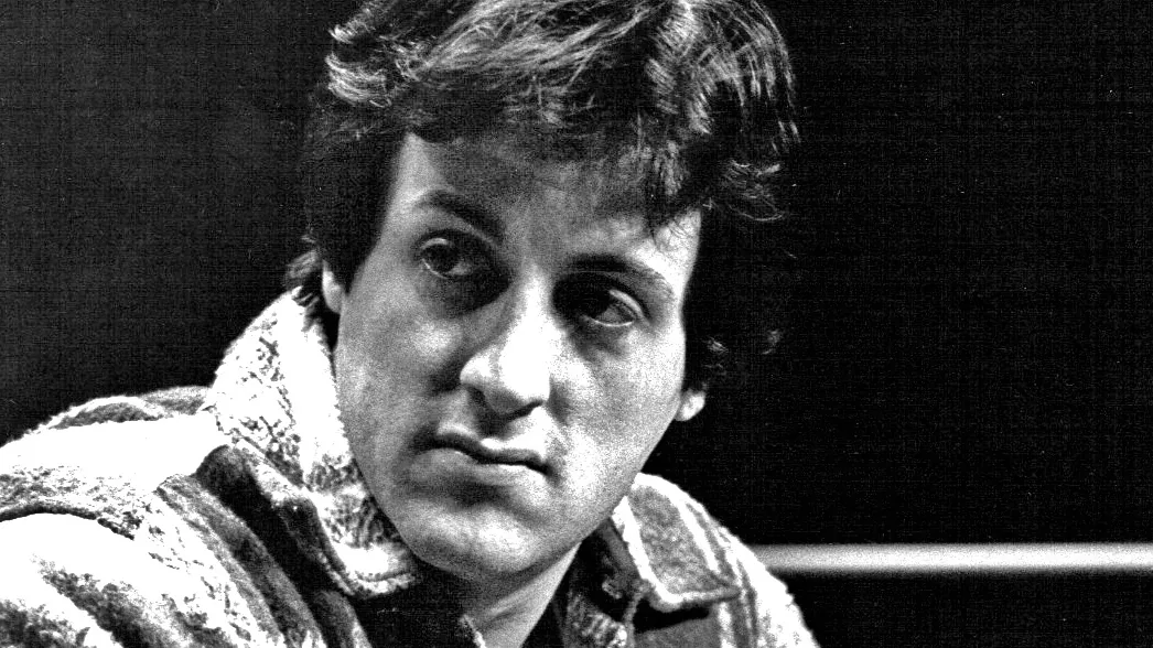 Young Sylvester Stallone at a boxing match in 1977, with thick dark hair worn long and a fringe falling over the forehead